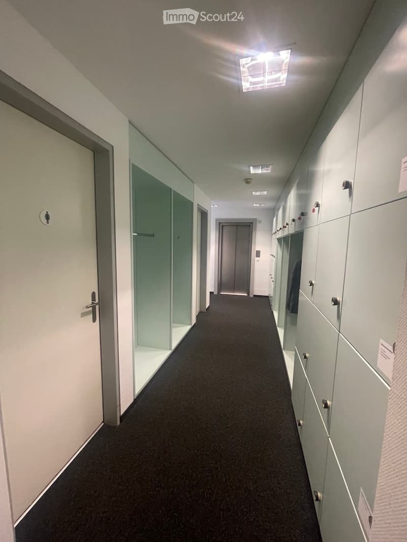 Empty hallway with white walls, black carpet, glass doors, lockers, and ceiling lights