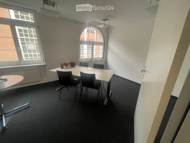 empty room with a long table, 4 chairs, 2 small tables with flowers, white walls, windows, black carpet
