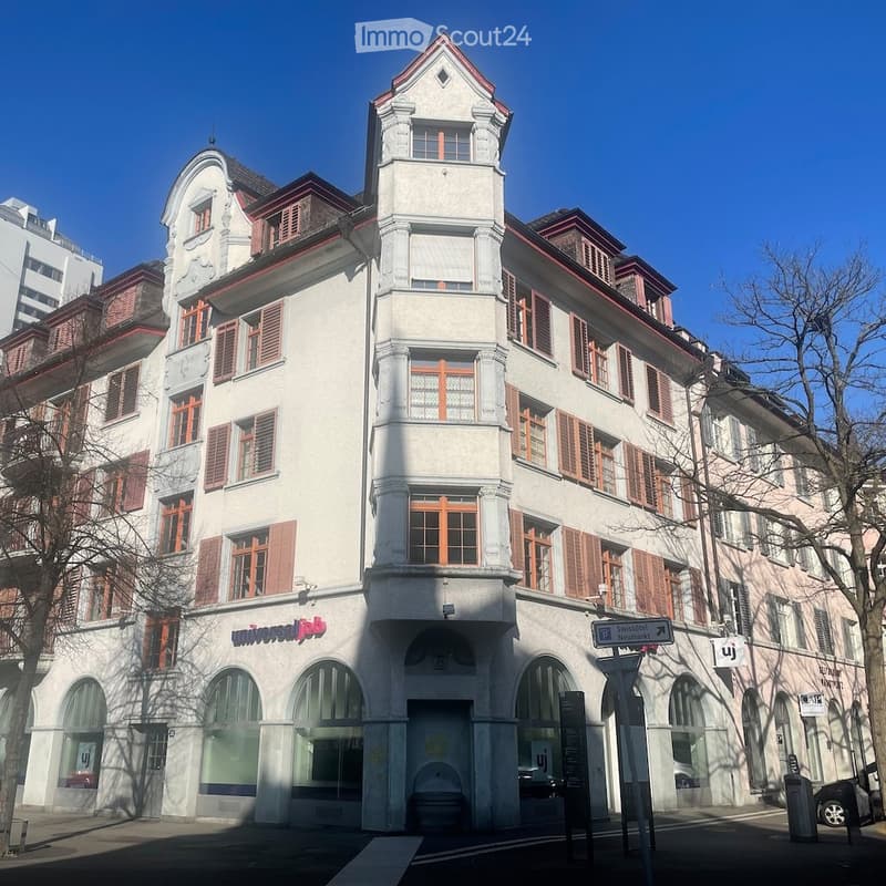 A multi-story building with white walls and red trim, arched windows, and a sign reading 'Universal Job'. The building has a modern architectural design with a combination of glass and metal elements. There are trees and a streetlight in front of the buil