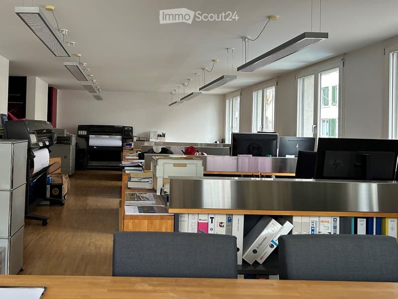 Office space with wooden floors, white walls, multiple desks, computers, printers, and storage boxes.