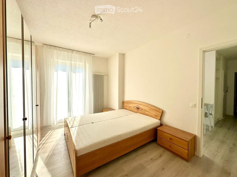A bedroom with a wooden bed, two nightstands, a wooden cabinet, and a white curtain. The room has a wooden floor and white walls. There is a white radiator on the wall and a ceiling lamp. The room has a glass door leading to another room with a white chai
