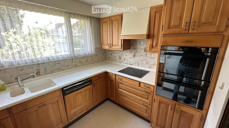 a fully fitted kitchen with wooden cabinets, white countertops, built-in stove, dishwasher, sink, double oven, window with curtains