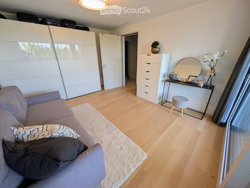 modern living room with gray couch, white rug, wooden floor, white walls, white cabinets, mirror, flowers, stool, and a view of the outside