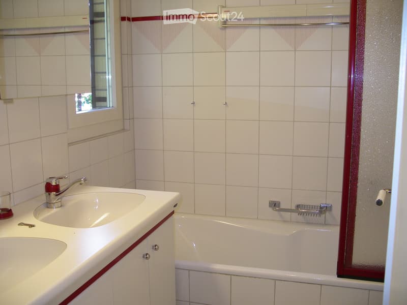 Bathroom with white tiles, double sink, bathtub, and a mirror. The sink has a faucet and a soap dish. The bathtub is white with a red border. The cabinet is white with red trim. A cup and a key are on the counter.