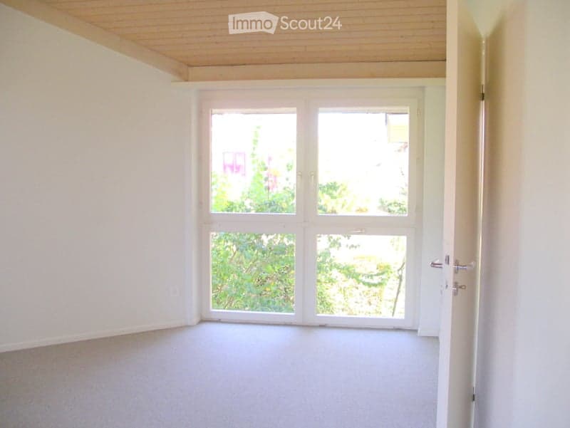 Empty room with white walls, wooden ceiling, and carpeted floor. Large windows with a view of trees outside.