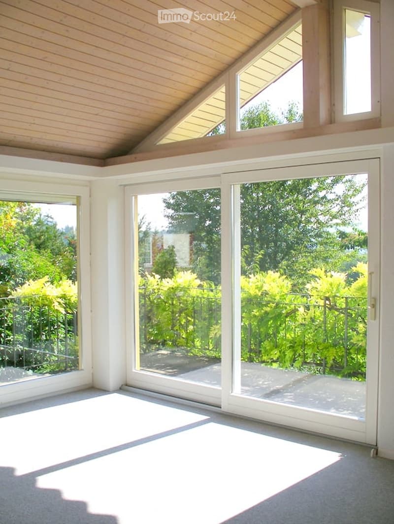 An empty room with white walls, a wooden ceiling, and large glass sliding doors leading to a balcony. The room has a white floor and is surrounded by greenery outside.