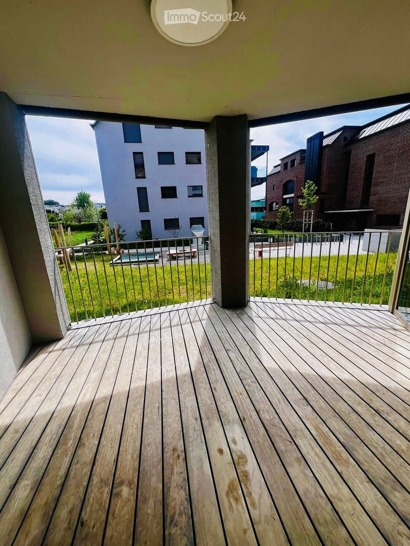 Empty balcony with wooden floor and metal railings, overlooking a grassy area with a small pool and buildings in the background.