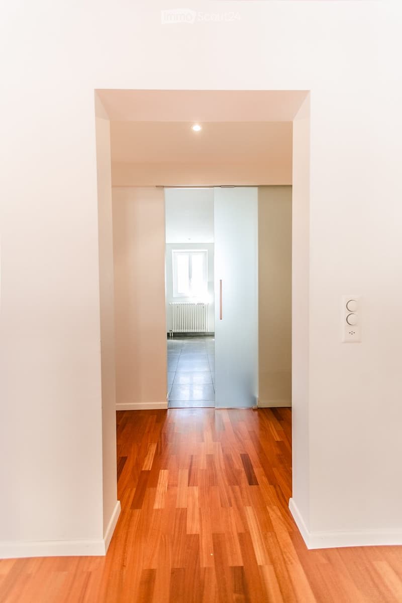 Empty hallway with wooden floor, white walls, sliding glass door, and a window with a radiator.