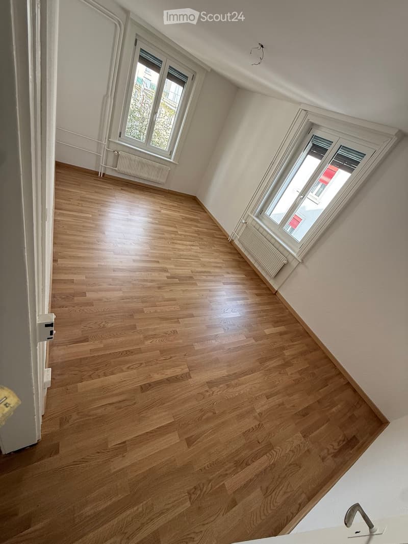 Empty room with wooden floors, white walls, and two windows with white frames. There are two white radiators under the windows.