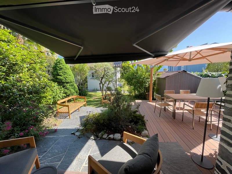 Wooden deck with outdoor dining table, chairs, wooden bench, flower plants, outdoor umbrellas, wall lamp, rock garden, pathway, building, houses, plants, garden wall