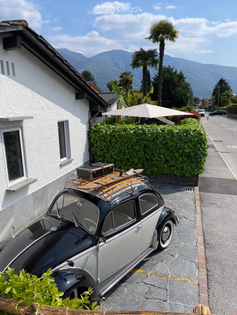 House exterior, vintage car parked in front, garden, trees, mountains in the distance