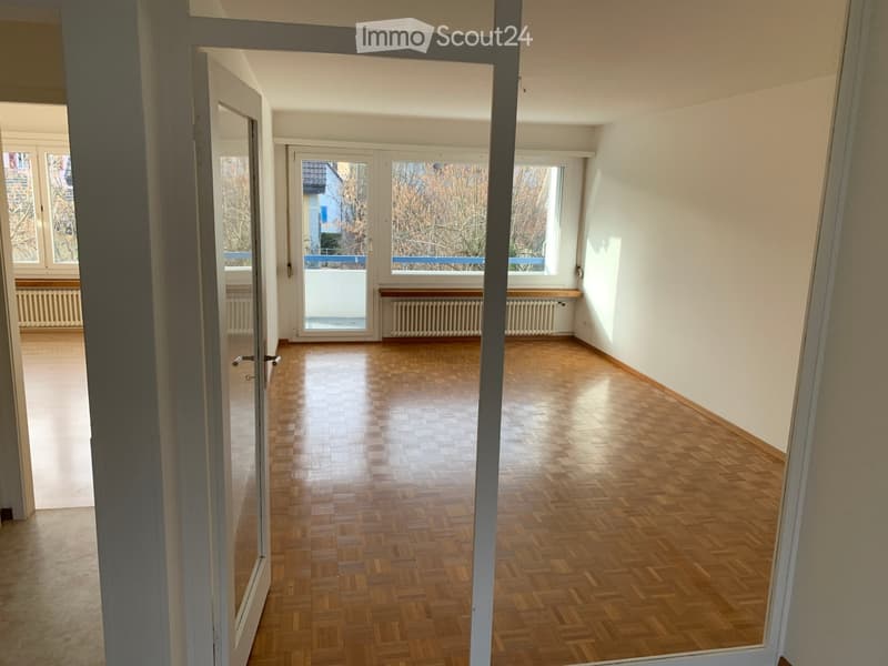 Empty room with wooden parquet flooring, white walls, and large windows with a balcony. There are two white radiators under the windows.