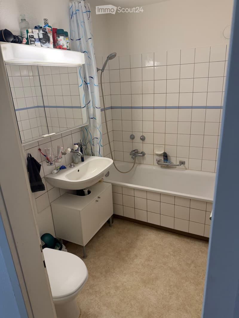 Bathroom with white tiled walls, a sink with a mirror, a bathtub with a shower curtain, a toilet, and a small cabinet under the sink.