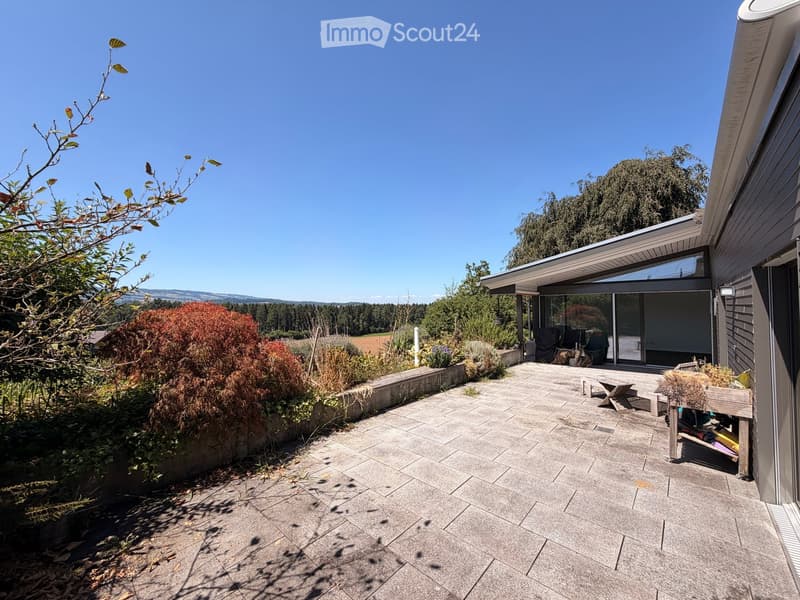 Outdoor patio area with stone tiles, wooden furniture, and surrounding greenery, featuring a modern house with large windows and a covered section.