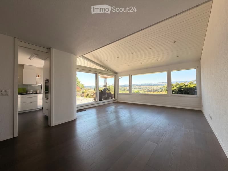 Bright, open room with dark wooden flooring, large windows, and a view of the landscape. The room features a modern kitchen area with white cabinets and stainless steel appliances visible in the background.