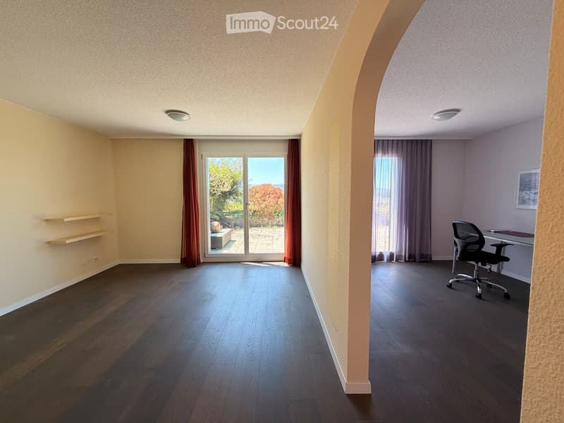 Open-plan room with wooden flooring, large sliding glass doors leading to a view, a desk with a chair, and built-in shelves.