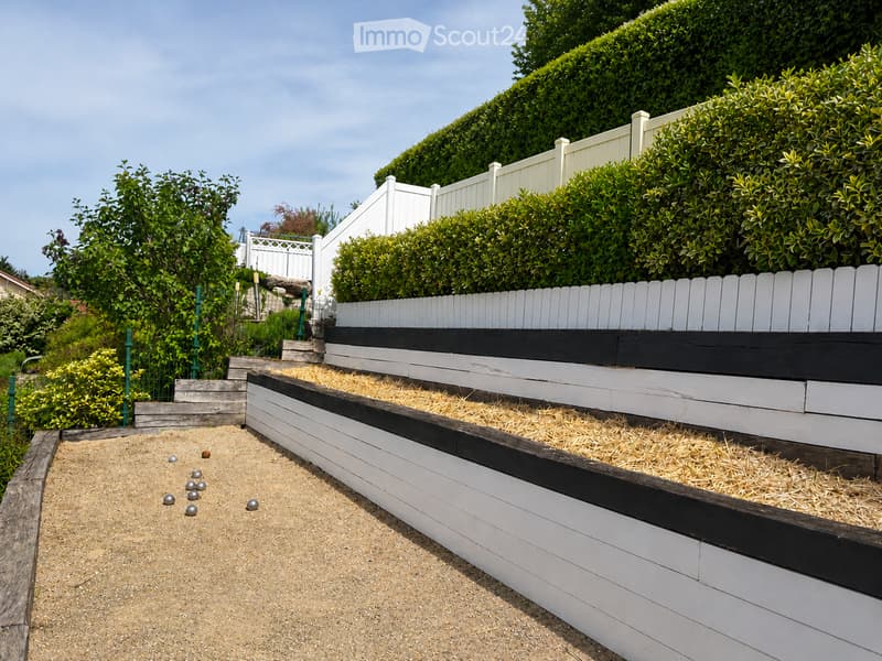 Garden / Outdoor Area. Outdoor area with a white wooden fence, bushes, and a staircase leading to a petanque court with a sandy surface and boules.