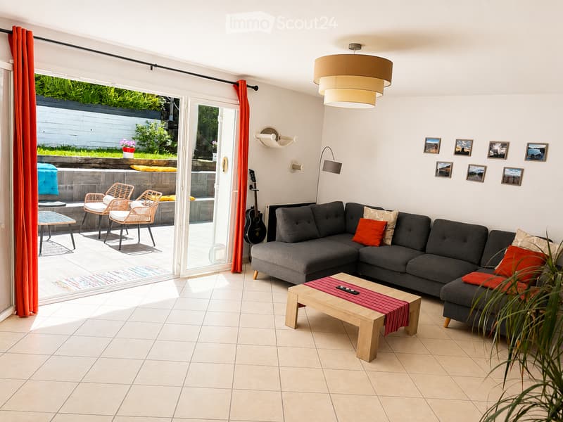 Modern living room with a grey sectional sofa, wooden coffee table, wicker chairs, and a view of a garden through large glass doors.