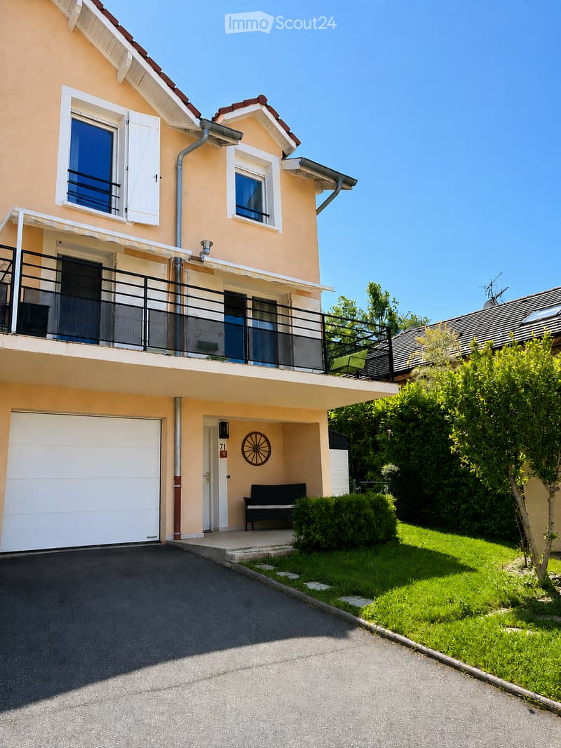 Two-story house with beige exterior, white-framed windows, garage, balcony, and small garden area.