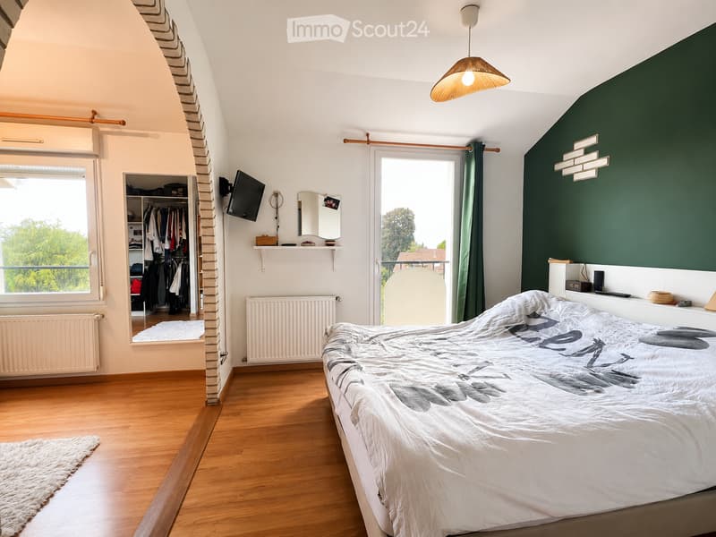 Cozy bedroom with wooden flooring, slanted ceiling, and large window. Features a double bed with a patterned duvet, a wardrobe with hanging clothes, a wall-mounted TV, a mirror, and a hanging lamp.