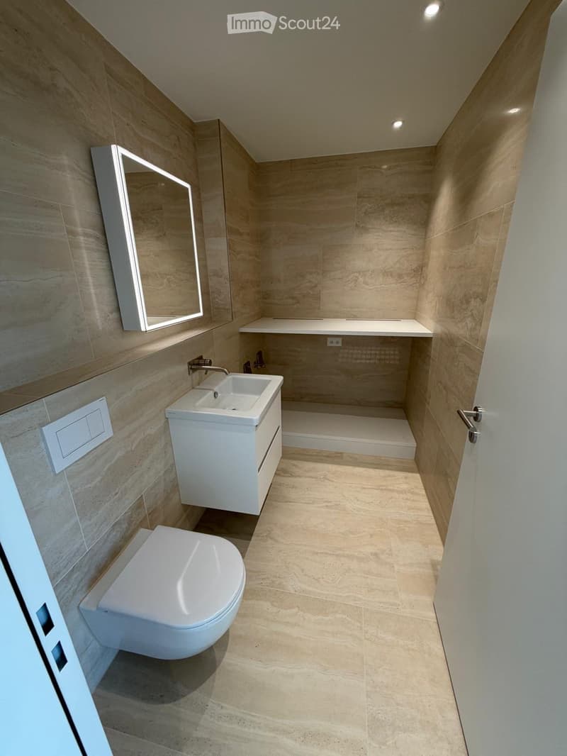 Modern bathroom with beige tiles, white toilet, sink, mirror, and power outlet. The shower area is partially visible.