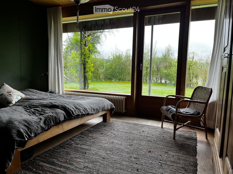 Bedroom with a bed, pillows, chair, rug, lamp, and radiator. Large windows with curtains.