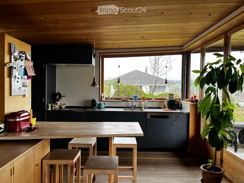 Open kitchen with wooden table, stools, and appliances. Large window with view of exterior.
