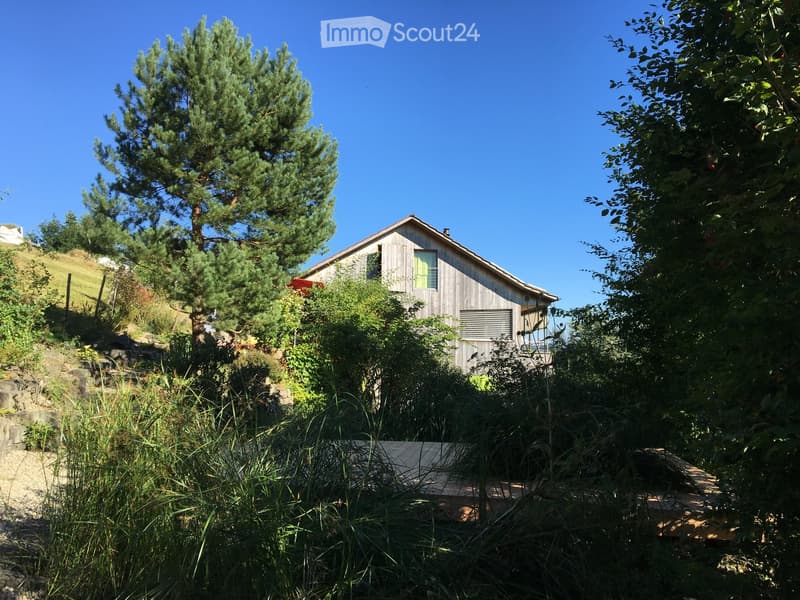 A wooden house with a red roof, nestled among trees and greenery on a hillside.