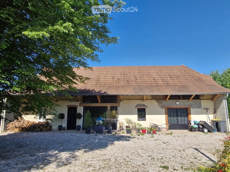 House. Two-story house with a tiled roof, beige walls, and a covered entrance area. The house features several windows, a black front door, and a gravel driveway. Potted plants and outdoor furniture are placed near the entrance, and a stack of firewood is