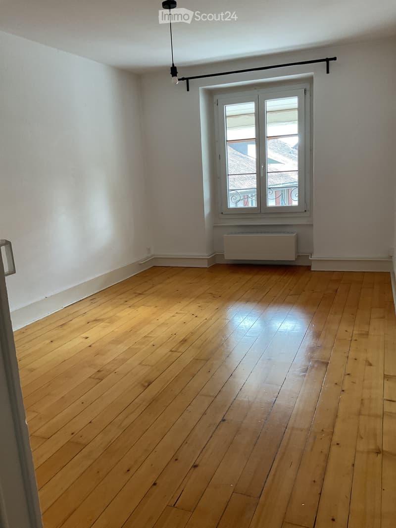 Unfurnished Room. Empty room with wooden flooring, white walls, and a large window with a view of rooftops. A radiator is visible beneath the window, and a single pendant light hangs from the ceiling.
