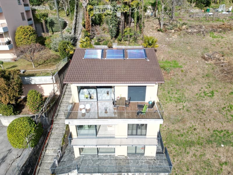 Two story house, beige exterior, brown roof, solar panels on roof, multiple balconies, fenced yard, neighboring buildings, greenery