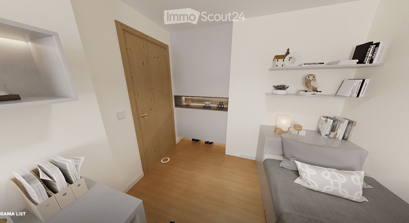 A modern, compact living space with a grey sofa, decorative pillows, built-in shelves holding books and decorative items, a small table with a lamp, and a small kitchen area visible through an open doorway.