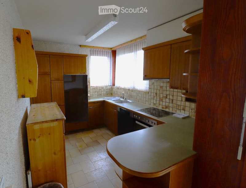 A small kitchen with wooden cabinets, a refrigerator, an oven, a sink, and a countertop. The space is well-lit with natural light from a window and has tiled flooring.