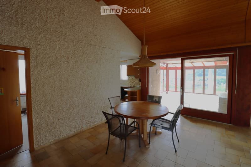 Dining area with a round wooden table and four chairs, textured wall, wooden ceiling, large window with view to outdoor area.