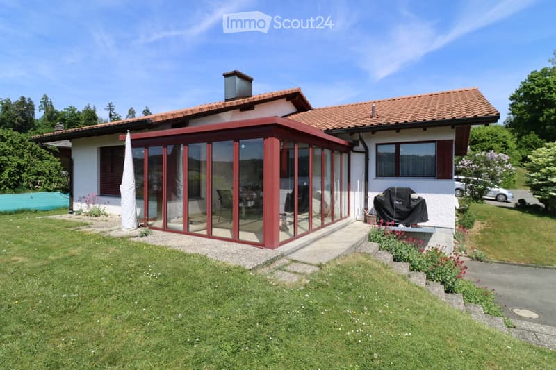 Two-story house with red-tiled roof, white walls, and a large glass-enclosed patio area. The patio has a few chairs and a table, and the surrounding yard is well-maintained with grass and flower beds.