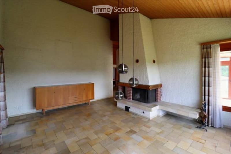 Living room with a fireplace, wooden cabinet, pendant lights, and a bench. The room features tiled flooring and large windows with curtains.