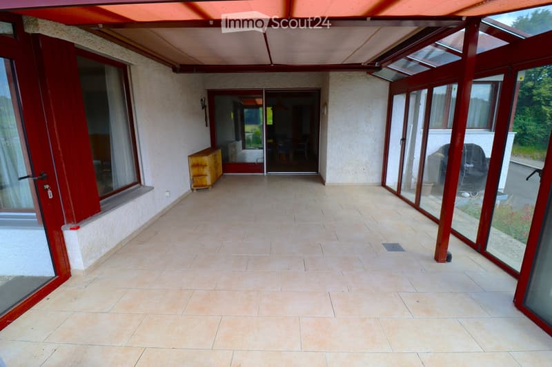 Spacious covered balcony with tiled flooring, large glass doors leading to the interior, and a small wooden cabinet near the entrance.