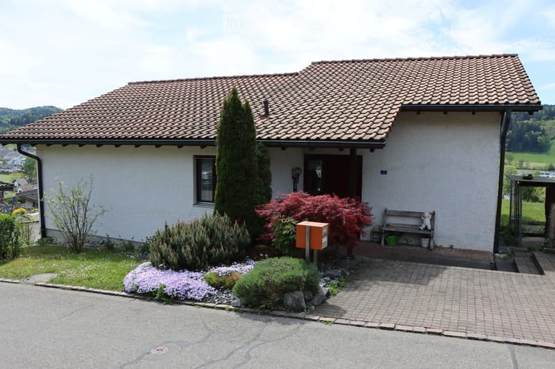 Single-story house with a tiled roof, white exterior walls, and a small garden with various plants and flowers. A wooden bench and a small table are placed near the entrance.