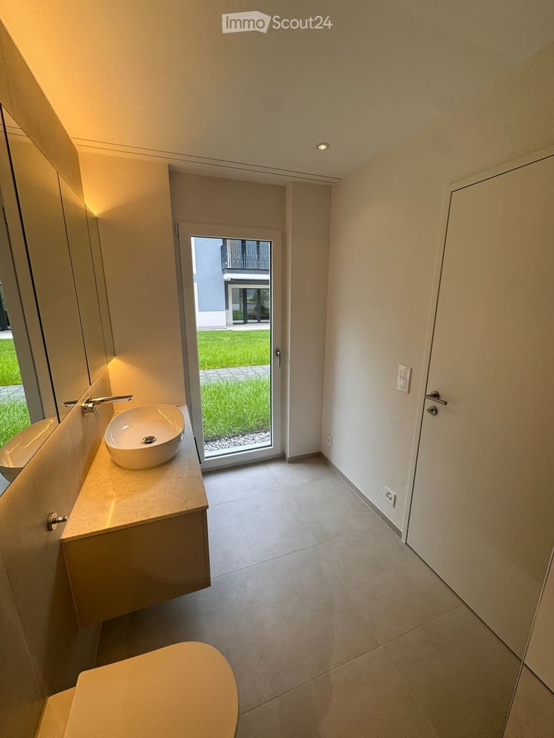 Modern bathroom with a white sink, toilet, and large window leading to a grassy area.