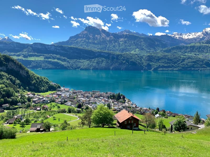 A picturesque village nestled by a serene lake, surrounded by lush green hills and towering mountains under a clear blue sky.