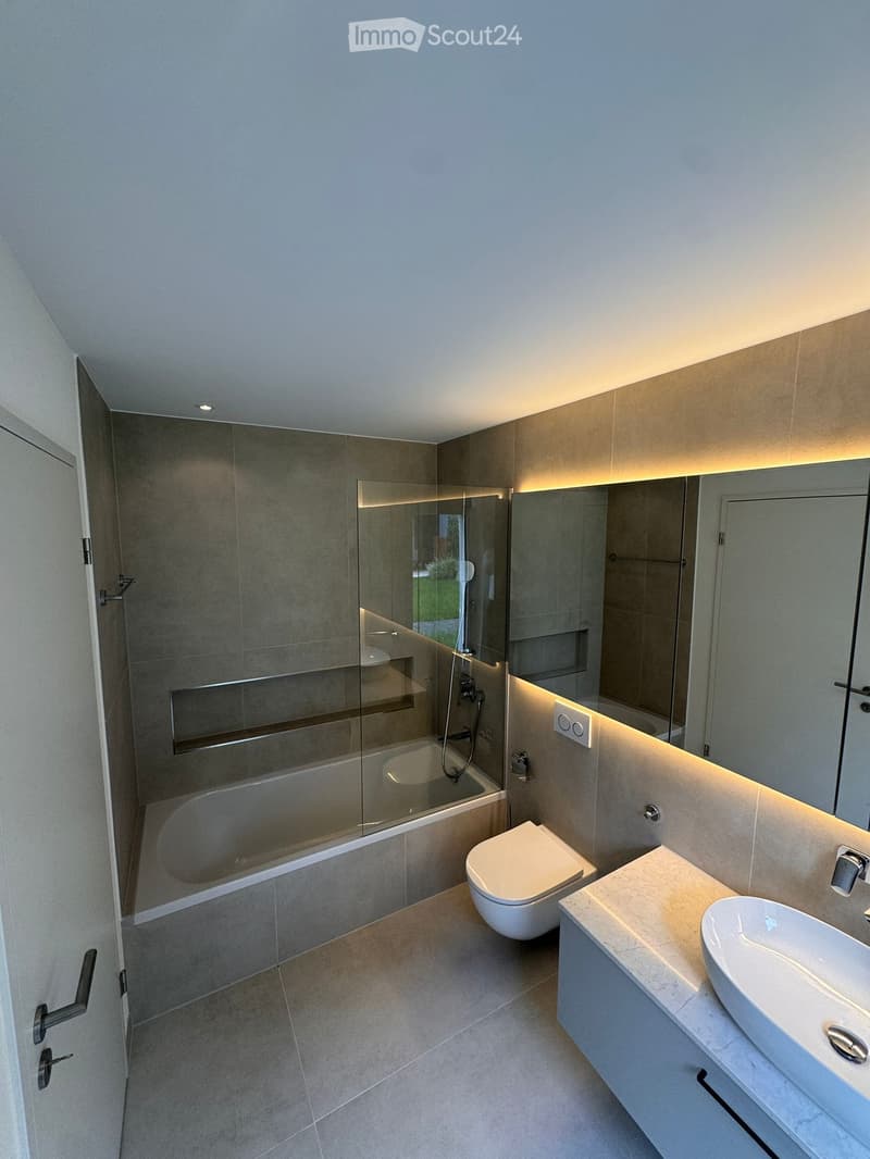 Modern bathroom with a bathtub, toilet, sink, and large mirror. Features include a glass-enclosed shower area and built-in lighting along the walls.