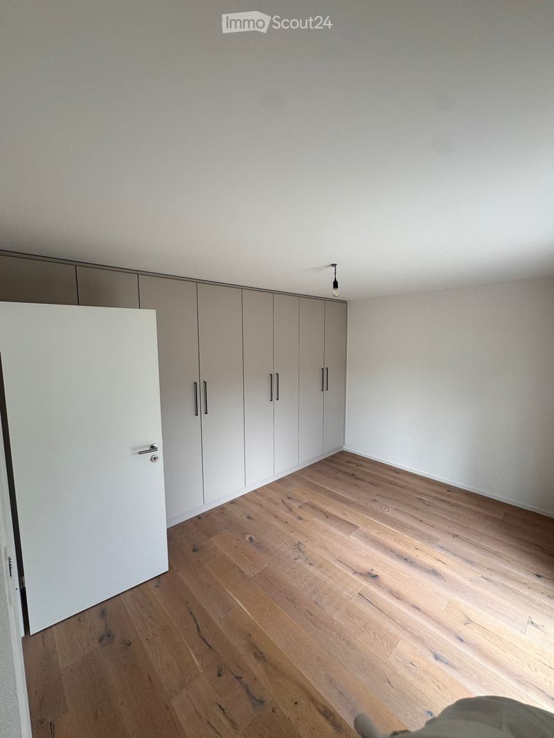 Empty room with light-colored wooden flooring and white built-in wardrobes along one wall. The room has a single ceiling light fixture and a door visible on the left side.