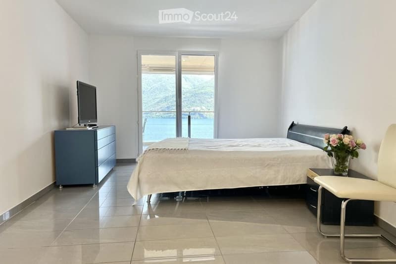 a neatly arranged bedroom with white tiled floor, a large bed, bedside table, flower vase, and a cabinet. The sliding glass door leads to a balcony overlooking the sea.