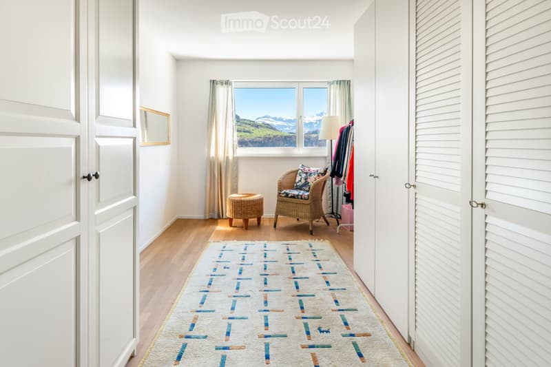 Furnished Room. Bright room with wooden flooring, large window offering a scenic mountain view, a wicker chair with a cushion, a small wicker stool, a patterned rug, a floor lamp, and a wardrobe with hanging clothes.