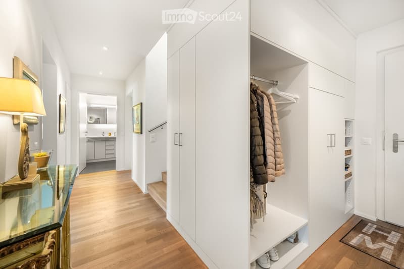 Corridor / Hallway. Modern hallway with light wooden flooring, white walls, built-in closet with hanging space, and a small glass table with a lamp and decorative bowl. A staircase leads to an upper level, and a bathroom is visible in the background.