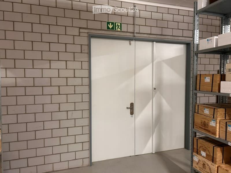 Corridor / Hallway. A hallway with a white double door and a green emergency exit sign above it. The walls are tiled with light-colored bricks, and there is a metal shelving unit with boxes on the right side.