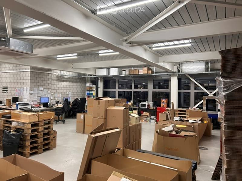 Furnished Room. A spacious, industrial-style office space with exposed metal beams and fluorescent lighting. The room is filled with desks, chairs, computers, and numerous cardboard boxes stacked on pallets, indicating an active workspace or storage area.