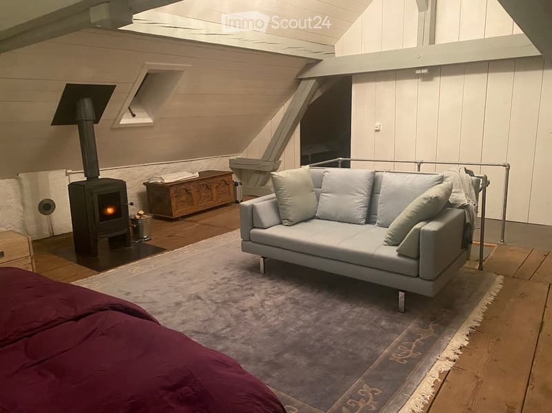 Furnished Room. Cozy attic living space with exposed wooden beams, a grey sofa with pillows, a wood stove, and a wooden cabinet.