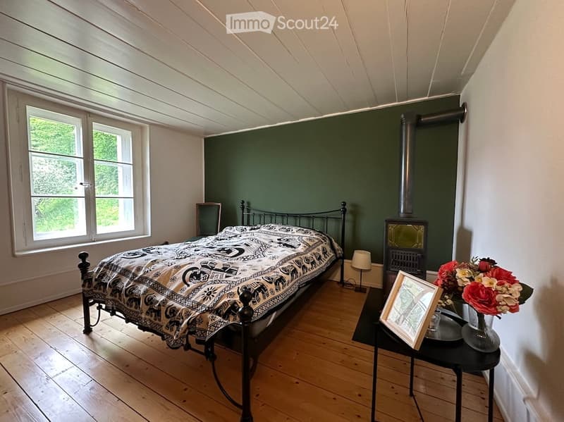 Bedroom. Cozy bedroom with wooden floors, slanted ceiling, green accent wall, fireplace, and large window. Features a patterned bedspread, nightstand with lamp, and decorative items.