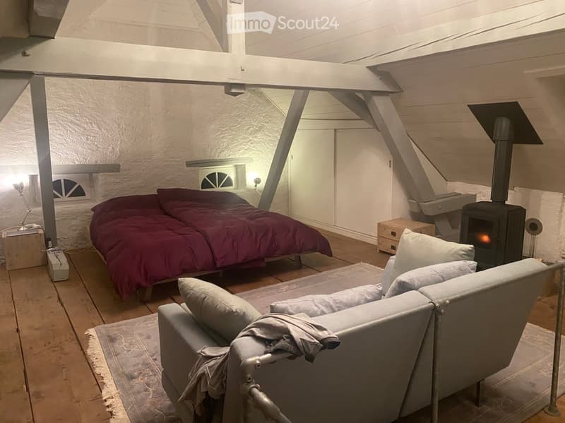 Furnished Room. Loft-style bedroom with exposed wooden beams, a double bed with red bedding, a grey sofa, a wood-burning stove, and a floor lamp.